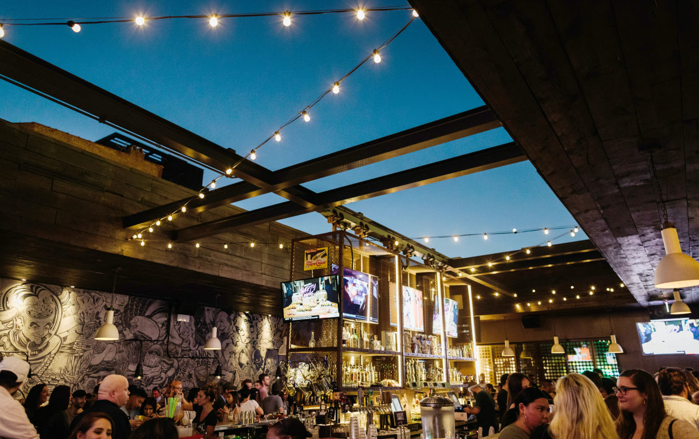 Busy urban rooftop bar patio at dusk with string lights and exposed steel beams, illuminated back bar with TVs and bottles, mural-covered wall and crowds enjoying drinks under a deep blue sky.