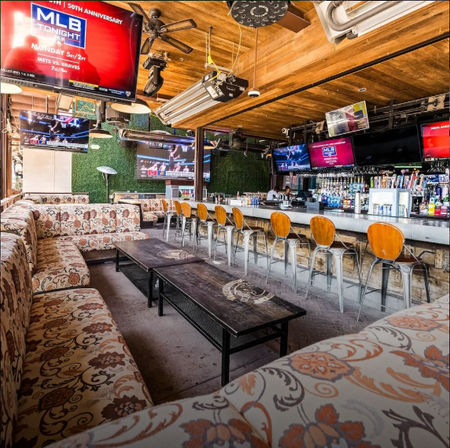 Vibrant urban sports-bar interior with patterned lounge sofas, long metal bar with wooden stools, multiple TVs showing MLB, wood ceiling and green wall