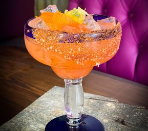 Bright orange margarita cocktail in a cobalt-rimmed goblet with crushed ice, an orange wedge and zesty chili-salt rim on a wooden table