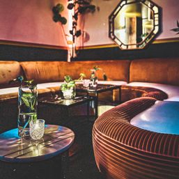 Cozy, dimly lit lounge seating with a curved velvet banquette, marble side tables, glass vases with green stems, candle and decorative wall mirror — moody retro-chic interior.