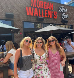 Three friends in summer dresses and sunglasses smiling on a sunny downtown outdoor bar patio beneath a red neon sign and black umbrellas