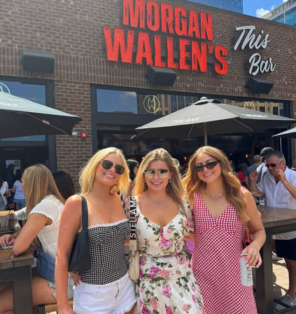 Three friends in summer dresses and sunglasses smiling on a sunny downtown outdoor bar patio beneath a red neon sign and black umbrellas