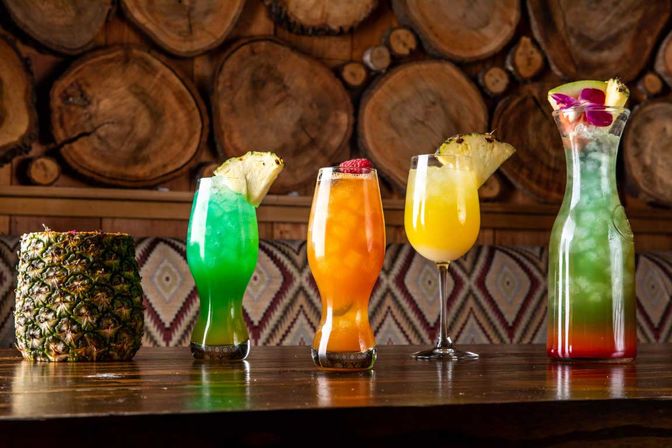 Row of vibrant tropical cocktails (green, orange, yellow) garnished with pineapple and orchid on a wooden bar in a tiki-style setting with stacked log wall backdrop