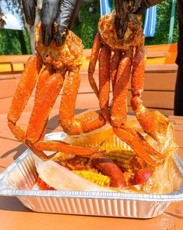 Gloved hands holding two spicy-seasoned crab clusters above an aluminum tray of Cajun seafood boil with corn and sausage on an outdoor picnic table