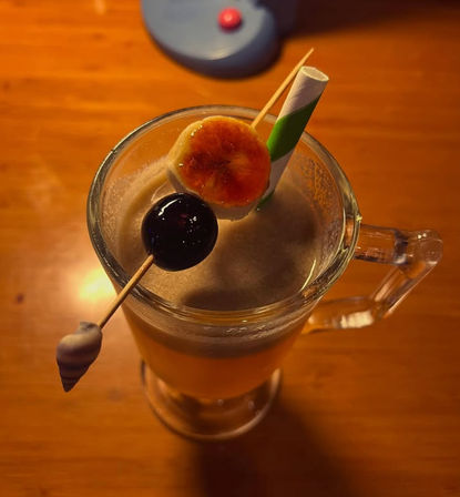 Frothy tropical cocktail in a clear glass mug on a wooden table, garnished with a caramelized banana slice and glossy cherry on a skewer plus a green-striped paper straw, warm amber lighting.