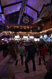 Lively crowd line-dancing on a wooden dance floor under a disco ball and twinkling string lights in a spacious indoor nightlife venue with mezzanine balcony.