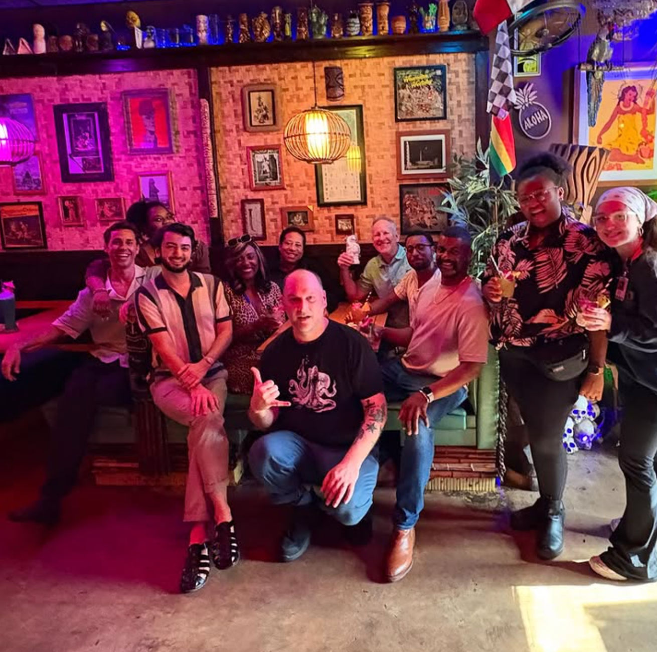 Smiling group posing with tropical cocktails in a colorful tiki-style bar lounge with rattan lights, framed art and lively nightlife vibe