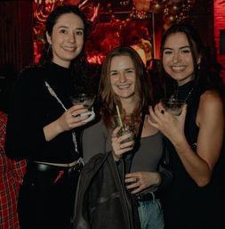 Three friends smiling and holding cocktails at a lively downtown bar with warm red lighting and balloon decorations in the background.
