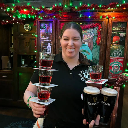 Playful bartender in a festive Irish pub balancing a tower of red shots on coasters and two pints of dark stout with creamy heads under holiday lights and tinsel