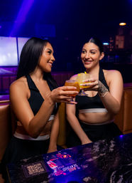 Two friends in black crop tops clinking fruity cocktails at a neon-lit cocktail bar booth, smiling under purple-blue nightclub lights with a QR menu on the marble table.