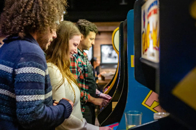 Three friends lined up at vintage arcade cabinets in a cozy arcade bar, smiling and playing retro video games.