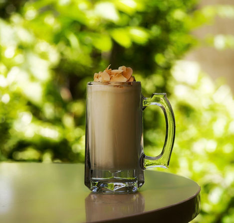 Frothy coconut latte topped with toasted coconut flakes in a clear glass mug on a sunlit outdoor table with a leafy green bokeh background