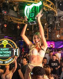 Lively nightclub scene: smiling dancer in pastel lingerie hoisted on shoulders, holding a glowing green champagne bottle trophy above a crowded VIP table with neon 'Happy Birthday' signage and party lights.