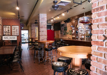 Empty cozy brick pub interior with wooden bar, round high-top tables, black padded bar stools, exposed brick columns, track lighting and framed wall photos