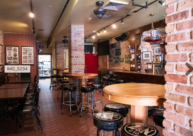 Empty cozy brick pub interior with wooden bar, round high-top tables, black padded bar stools, exposed brick columns, track lighting and framed wall photos