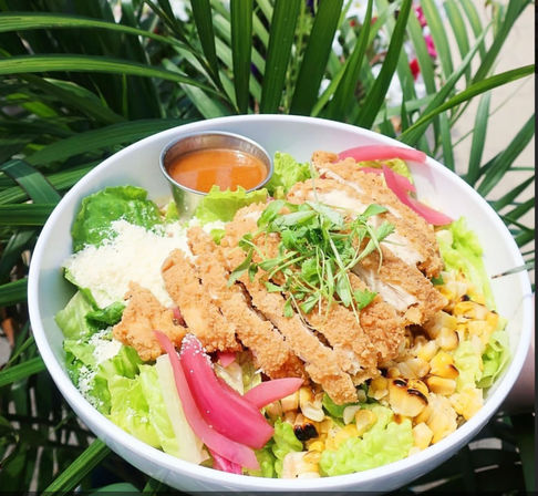 Crispy breaded chicken sliced over romaine salad with charred corn, pickled red onions, grated Parmesan and microgreens, with a metal cup of tangy dressing in a white bowl set against green tropical foliage.