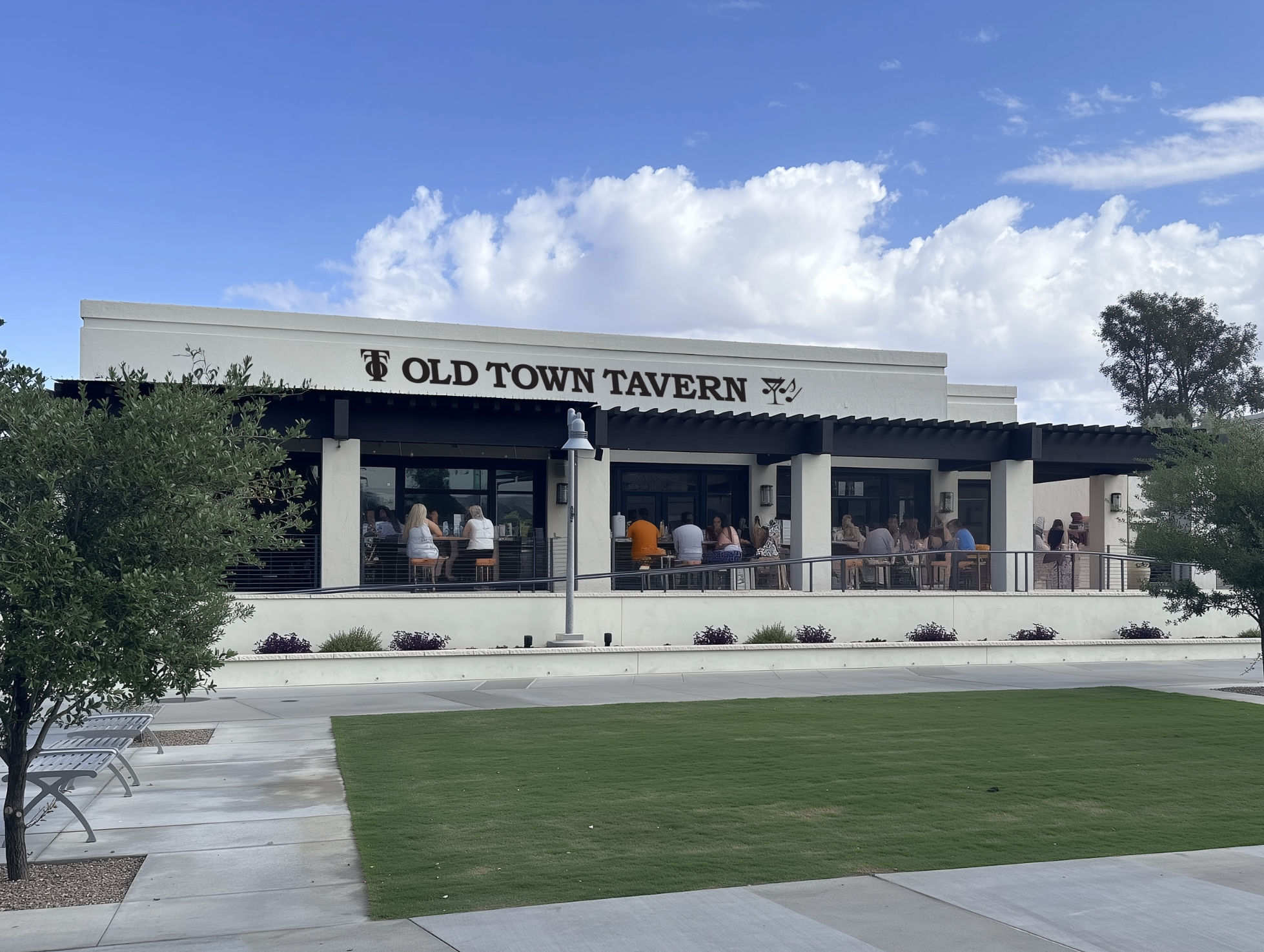 Sunny suburban eatery with white stucco facade and covered outdoor patio dining, patrons seated along the railing, manicured lawn and walkway in foreground under a blue sky