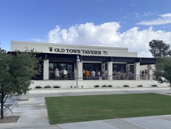 Sunny suburban eatery with white stucco facade and covered outdoor patio dining, patrons seated along the railing, manicured lawn and walkway in foreground under a blue sky