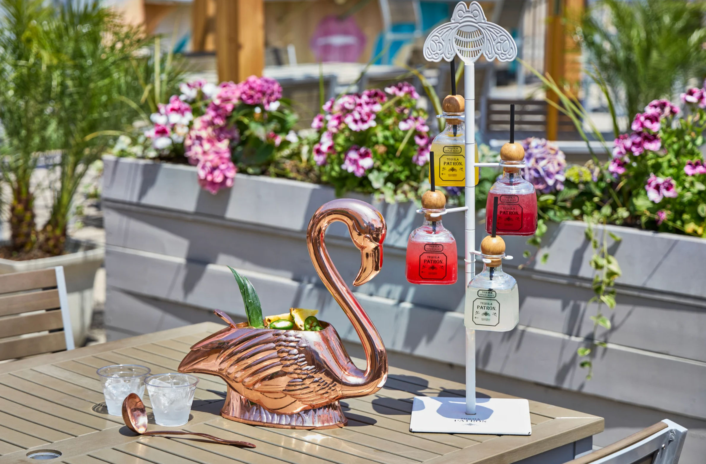 Shiny copper flamingo pitcher filled with tropical garnish beside a white stand of small colorful liquor bottles, two iced glasses and a spoon on a sunny outdoor patio table with pink flowering planters
