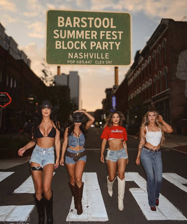 Four women in summer-festival outfits—crop tops, denim shorts, cowboy boots and hats—walking across a downtown Nashville crosswalk at dusk beneath a green Summer Fest Block Party sign.