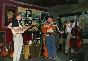 Live rockabilly trio on a cozy retro club stage: two guitarists (electric and acoustic) and an upright bassist playing amid drums, amps and vintage posters.