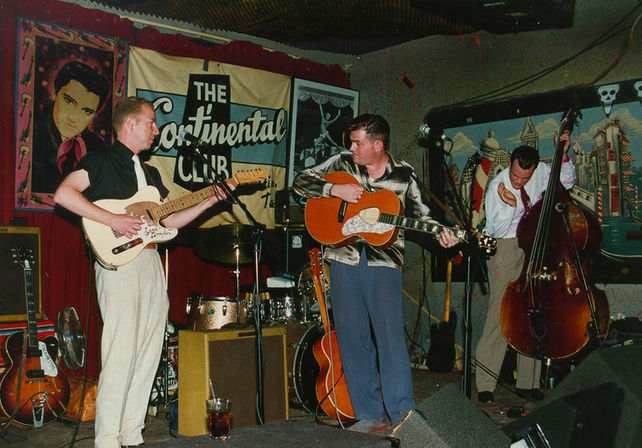 Live rockabilly trio on a cozy retro club stage: two guitarists (electric and acoustic) and an upright bassist playing amid drums, amps and vintage posters.