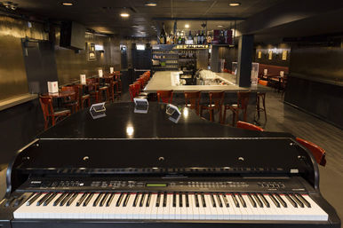 Dim, intimate piano bar interior with a black grand piano keyboard in the foreground, long central bar, wooden stools and bottles on back shelves.