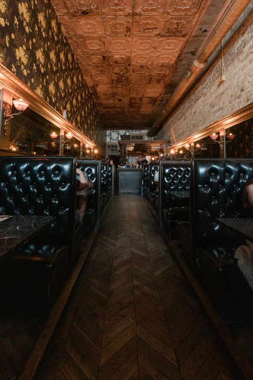 Narrow vintage restaurant interior with tufted black leather booths, chevron wood floor, patterned tin ceiling and warm wall sconces creating a cozy, dimly lit ambiance.