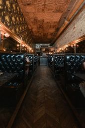 Narrow vintage restaurant interior with tufted black leather booths, chevron wood floor, patterned tin ceiling and warm wall sconces creating a cozy, dimly lit ambiance.