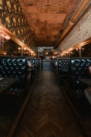 Narrow vintage restaurant interior with tufted black leather booths, chevron wood floor, patterned tin ceiling and warm wall sconces creating a cozy, dimly lit ambiance.