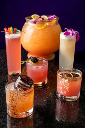 Colorful pink and orange cocktails on a glossy black bar counter: a large orange punch bowl garnished with lemon slices and edible flowers, tall frothy highballs with orchid and gummy skewers, and short rocks glasses topped with fruit skewers, dried citrus and a chili-salt rim.