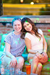 Two smiling women seated on an urban rooftop lounge sofa at night with colorful disco lights and a cocktail with glasses on a low table