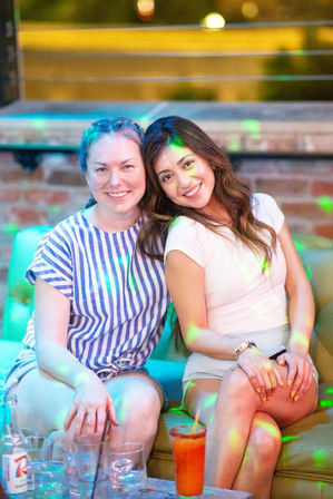 Two smiling women seated on an urban rooftop lounge sofa at night with colorful disco lights and a cocktail with glasses on a low table