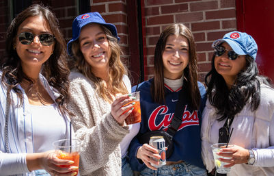 Four smiling young women in Chicago wearing Cubs hats and a Cubs jersey, holding cocktails and beer and posing outdoors by a brick wall