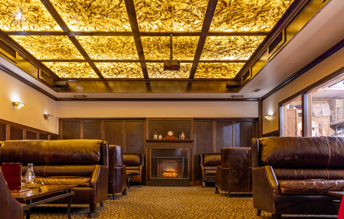 Warm, cozy lounge with dark leather sofas, patterned carpet, glowing fireplace and an amber backlit coffered ceiling