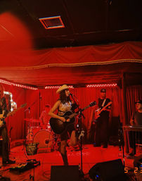 Lead country singer in a cowboy hat strums an acoustic guitar with her band on an intimate red-lit club stage — live music at a small venue with drummer, electric guitarist and vintage curtains.