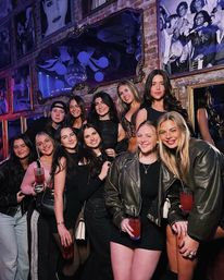 Group of young women on a night out at a brick-walled lounge with framed photos and a disco ball, holding red cocktails and smiling for a group photo.