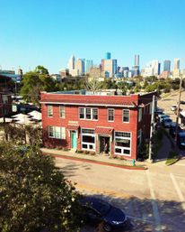 Aerial view of a red-brick two-story corner building with teal doors and tile awnings, leafy foreground, street with parked cars, and a distant downtown skyline under a clear blue sky.