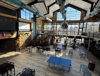 Spacious industrial-style indoor event space with vaulted skylights and exposed ductwork, mezzanine and wooden staircase, floor-to-ceiling windows, string lights and a large disco ball, scattered tables and a ping-pong table on light wood floors.