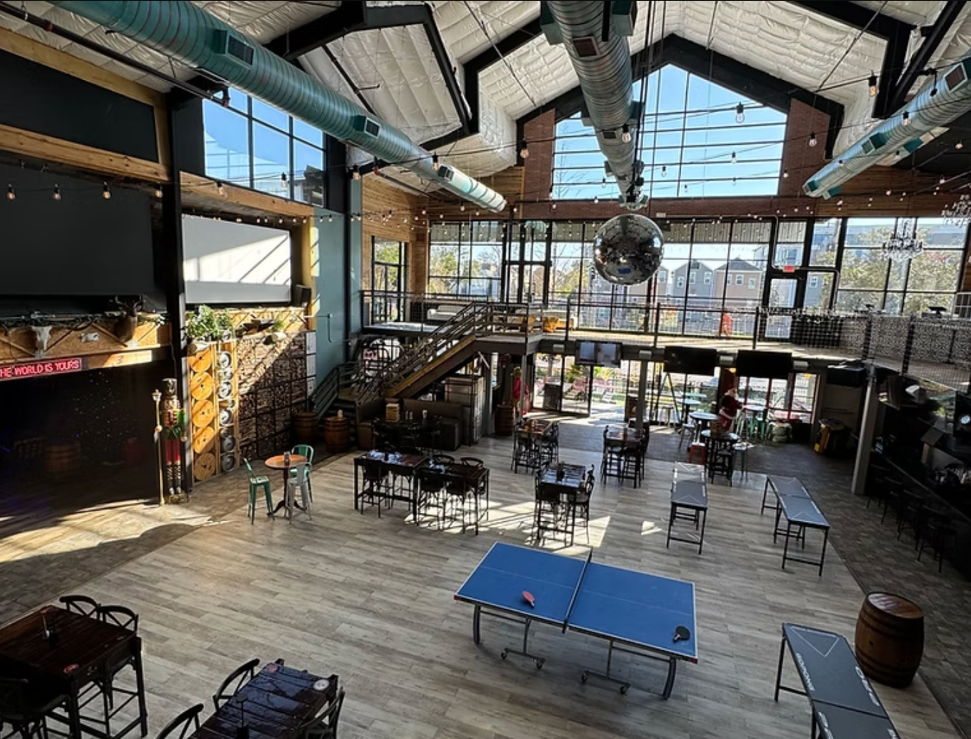 Spacious industrial-style indoor event space with vaulted skylights and exposed ductwork, mezzanine and wooden staircase, floor-to-ceiling windows, string lights and a large disco ball, scattered tables and a ping-pong table on light wood floors.