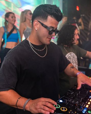 DJ in sunglasses and silver chains operating a colorful digital mixer in a neon-lit nightclub with dancers and a lively crowd in the background.