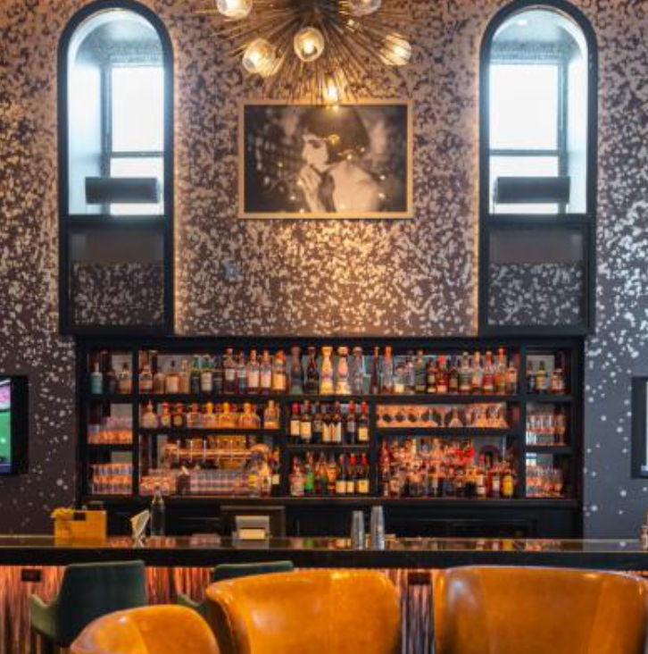 Chic urban cocktail bar interior with backlit liquor shelves, amber leather lounge chairs at a sleek bar, patterned metallic wallpaper, arched windows, a black-and-white portrait, and a starburst chandelier.