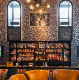 Chic urban cocktail bar interior with backlit liquor shelves, amber leather lounge chairs at a sleek bar, patterned metallic wallpaper, arched windows, a black-and-white portrait, and a starburst chandelier.