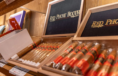 Open wooden cigar boxes on a shelf revealing neat rows of cellophane-wrapped cigars with red-and-black bands against a rustic wood backdrop.