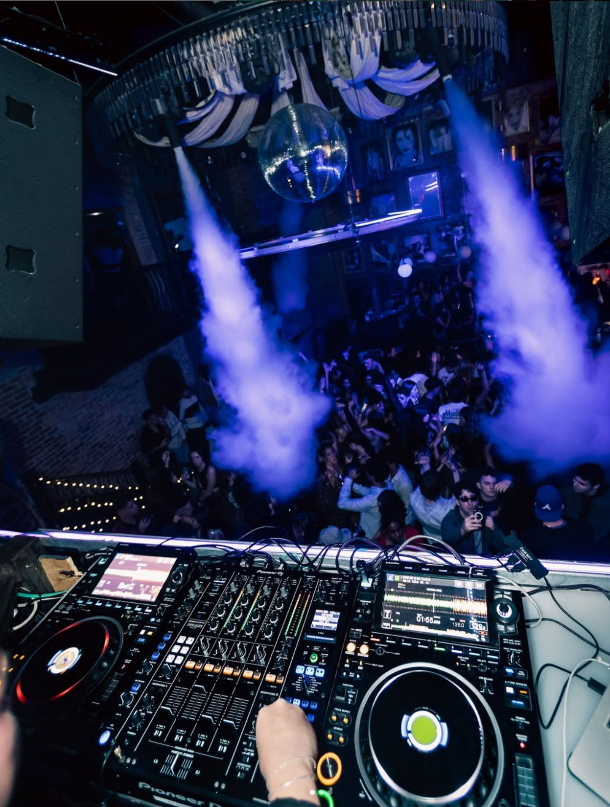 DJ booth overlooking a packed nightclub dance floor with blue lights, fog blasts and a mirrored disco ball — lively urban nightlife scene.