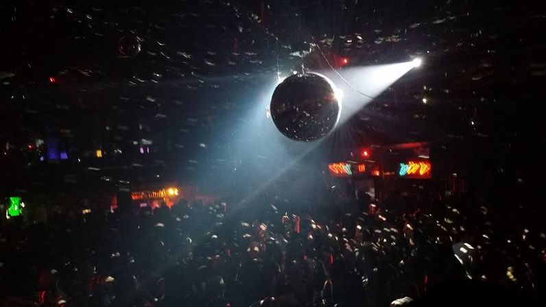 Packed nightclub dance floor with a mirrored disco ball lit by a bright white spotlight, scattering glittering light across the crowd and colorful bar lights in the background.