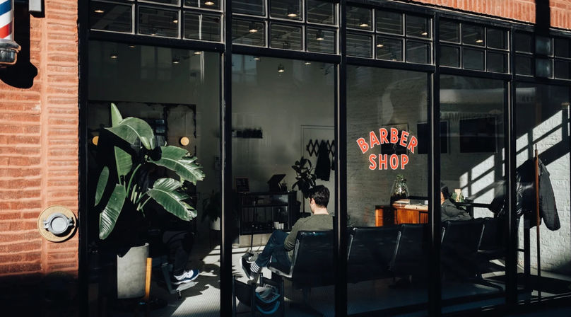 Sunlit urban barber shop storefront with glass windows, waiting chairs, seated customers and large potted plant