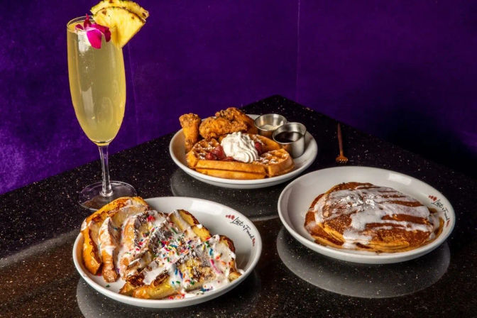 Brunch spread on a sparkly black table: a pineapple cocktail in a flute, crispy chicken and waffles with syrup and whipped cream, powdered-sugar pancakes, and sprinkles-topped French toast against a purple velvet backdrop.