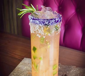 Tall pink citrus cocktail with ice, diced jalapeño and rosemary sprig, in a salt- and purple-rimmed glass on a wooden table against a magenta tufted booth.