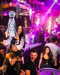 Smiling woman on a friend's shoulders holding an oversized bottle prop in a neon‑lit nightclub, lively crowd, sparklers and VIP bottle service vibes